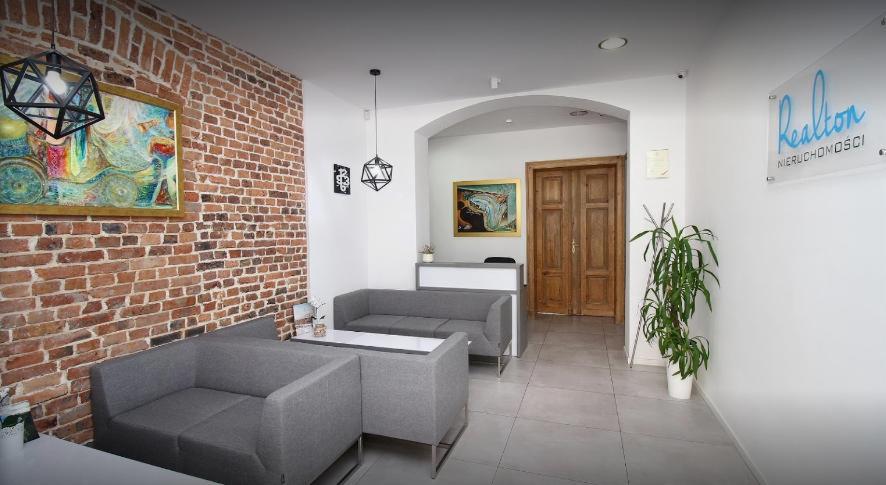 Wnętrze biura agencji nieruchomości z szarymi sofami, ceglaną ścianą z obrazem, recepcją w tle i logo firmy na ścianie.