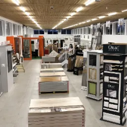 Wnętrze przestronnego salonu sprzedaży z ekspozycją materiałów wykończeniowych, w tym palet z panelami podłogowymi, ekspozytorów z roletami i listwami dekoracyjnymi, oraz fragmentów okien i drzwi...