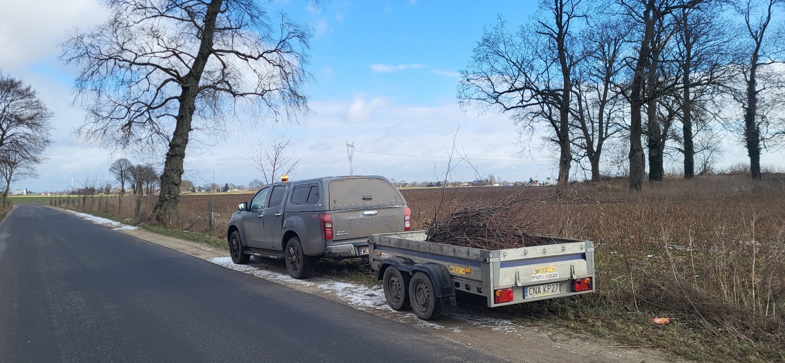 Szary pickup Isuzu z przyczepą pełną gałęzi, zaparkowany na poboczu drogi wiejskiej, w tle nagie drzewa i pole.