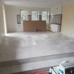 Układanie szarych płytek podłogowych w przestronnym, jasnym pomieszczeniu z trzema oknami zabezpieczonymi folią, widoczna maszyna do cięcia płytek na pierwszym planie i puszki z farbą w tle.