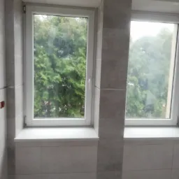 Dwa okna w łazience z jasnoszarymi płytkami, widok na zieleń za oknem, fragment instalacji elektrycznej po lewej stronie.