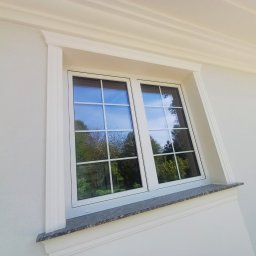 Firma Budowlana JackBud Jacek Kalinowski - Biała elewacja z oknem w białej ramie, ozdobnymi listwami i kamiennym parapetem. W szybach odbija się zieleń drzew.