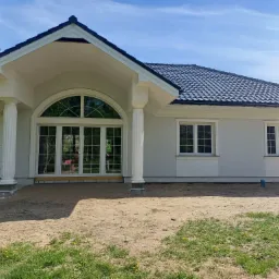 Elegancki, parterowy dom z jasną elewacją, ciemną dachówką i kolumnami przy wejściu. Duże, łukowe okno. Widoczne detale sztukaterii i świeżo zagospodarowany teren.