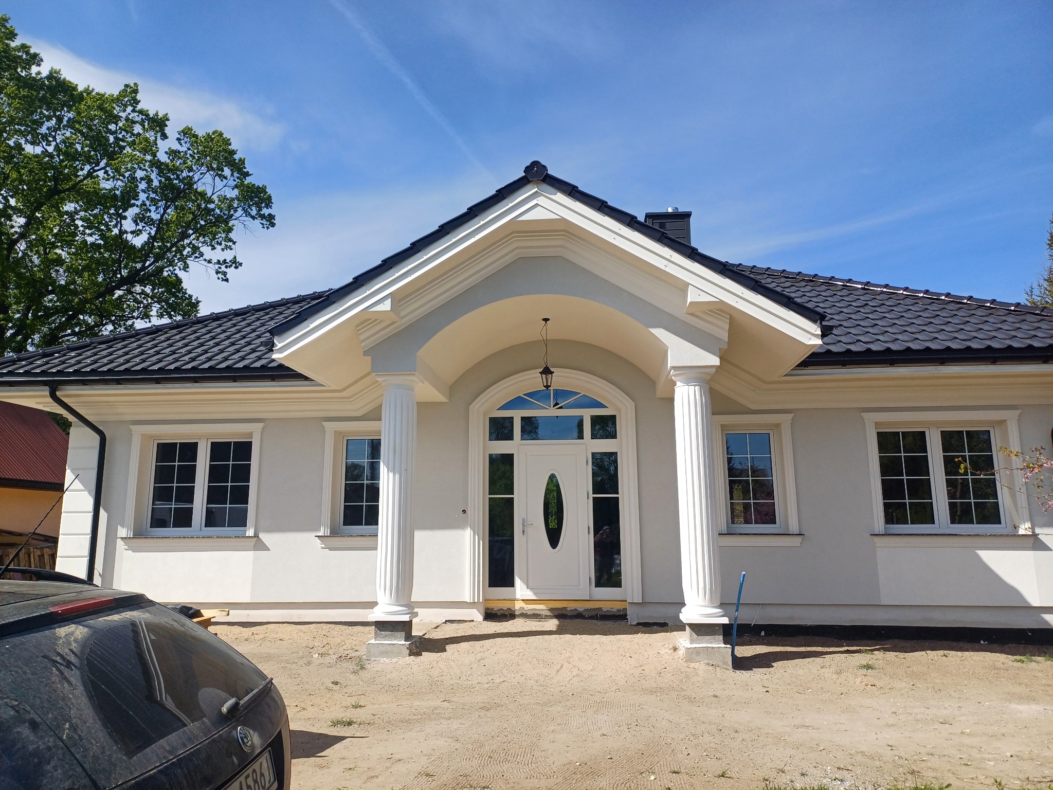Biały dom jednorodzinny z kolumnami przy wejściu, czarny dach, okna z białymi ramami, samochód częściowo widoczny z lewej strony. Błękitne niebo.