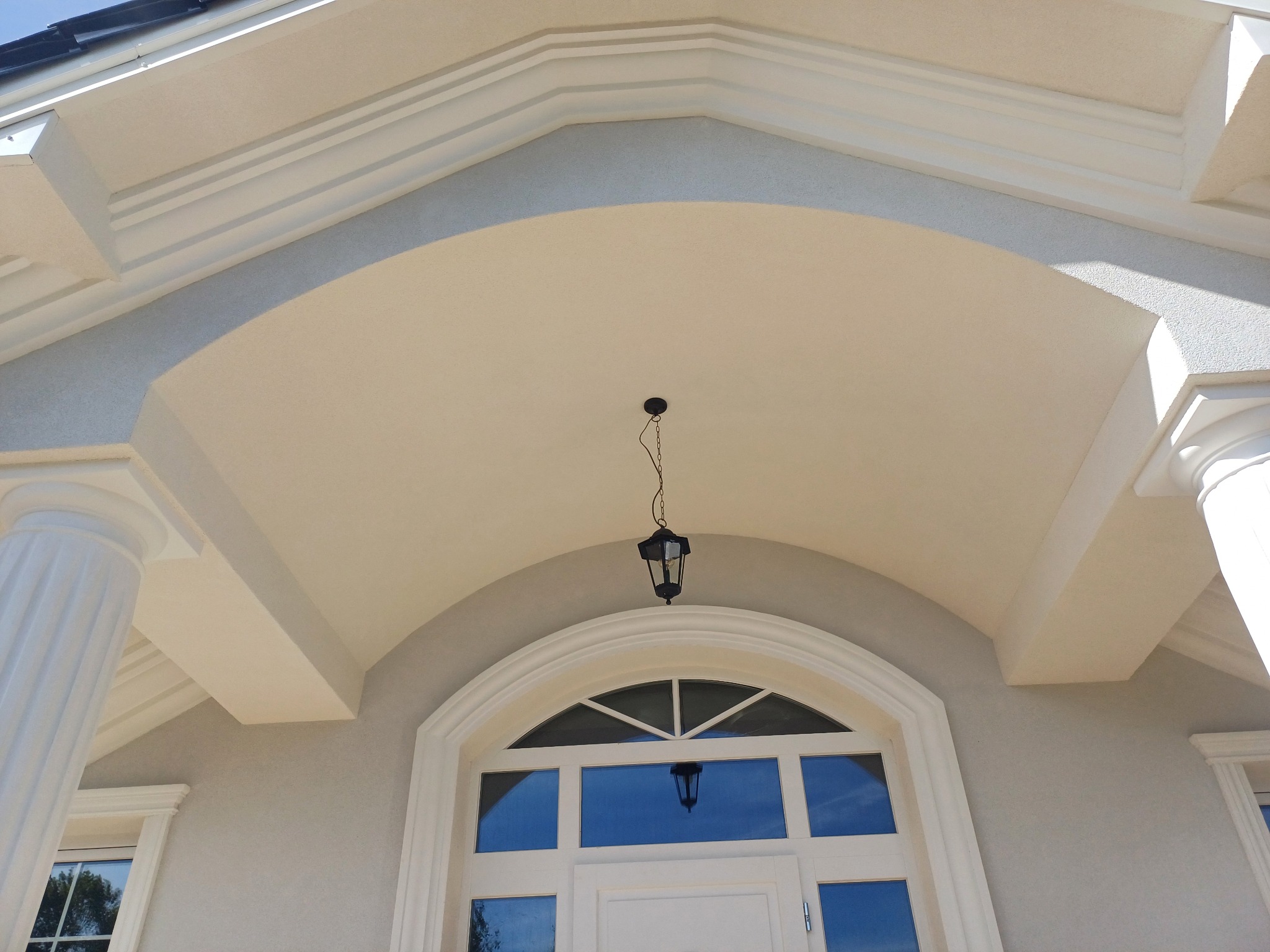 Eleganckie wejście do budynku z kolumnami i łukowym oknem, oświetlone wiszącą lampą. Detale architektoniczne podkreślone jasną kolorystyką.