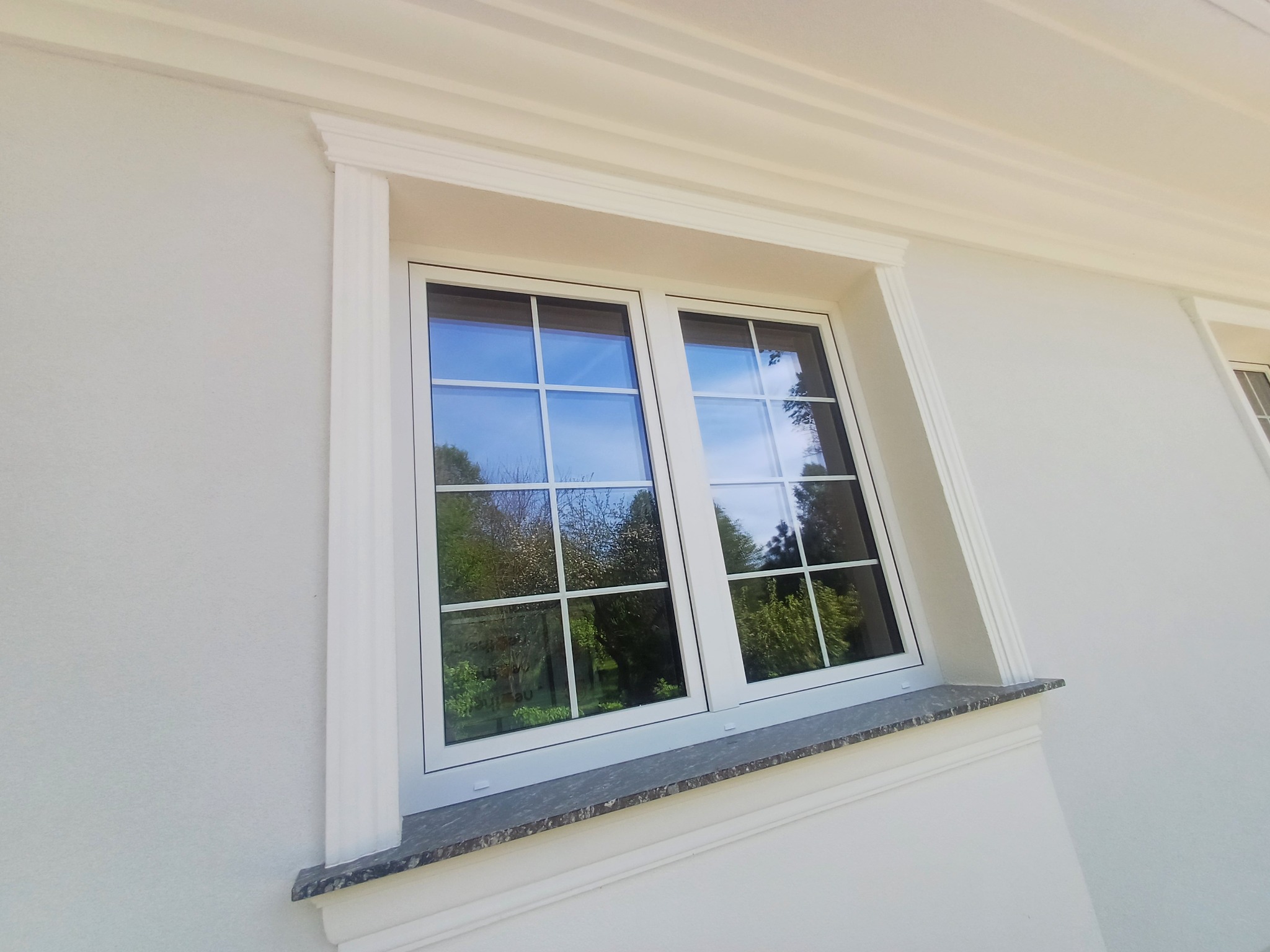 Biała elewacja z oknem w białej ramie, ozdobnymi listwami i kamiennym parapetem. W szybach odbija się zieleń drzew.