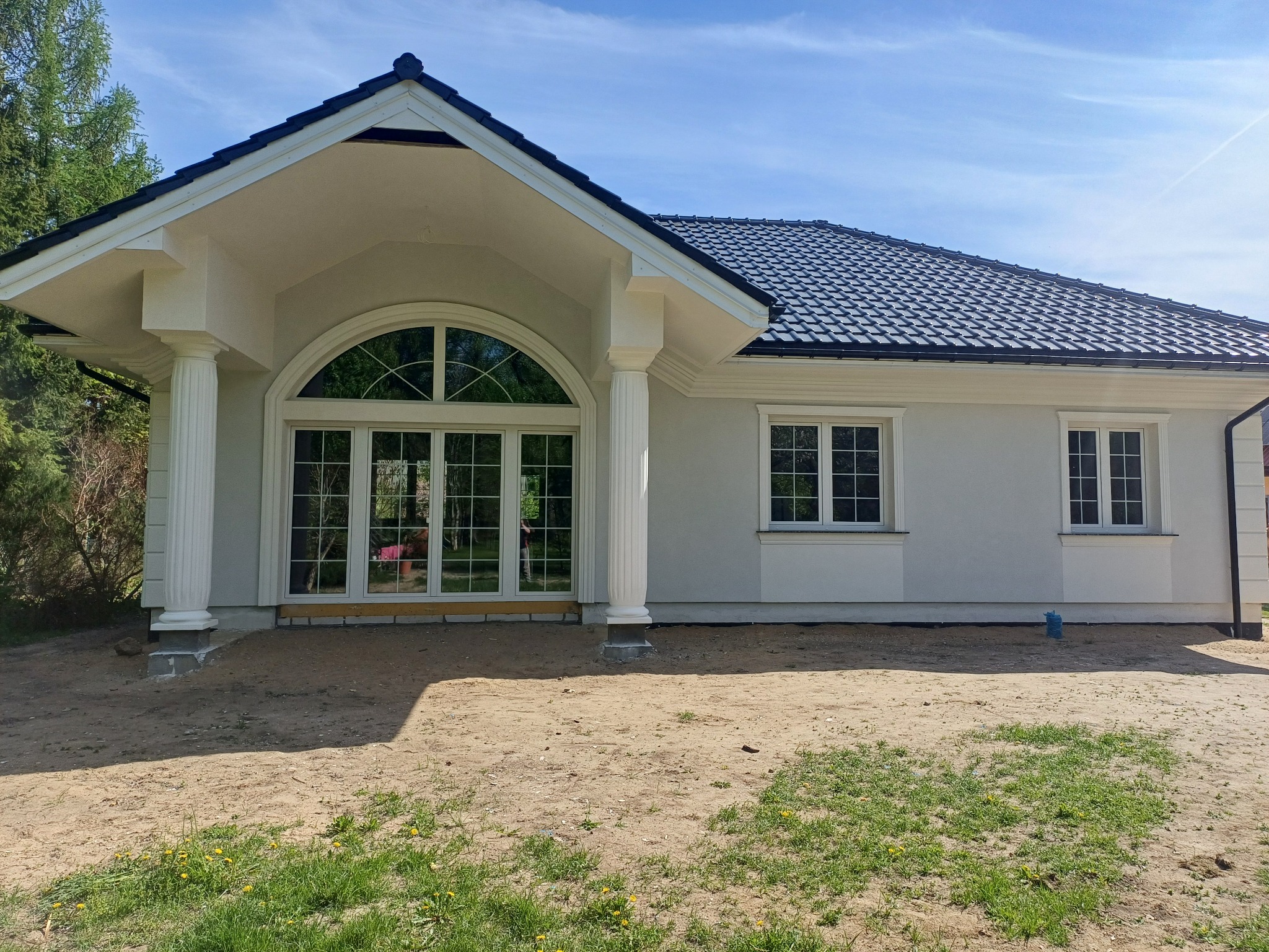 Elegancki, parterowy dom z jasną elewacją, ciemną dachówką i kolumnami przy wejściu. Duże, łukowe okno. Widoczne detale sztukaterii i świeżo zagospodarowany teren.