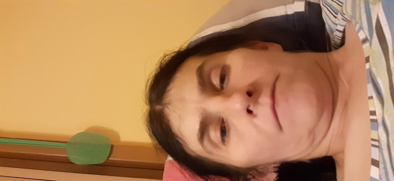 Portret kobiety leżącej na łóżku z zieloną packą na muchy w tle, skierowana twarzą do góry.