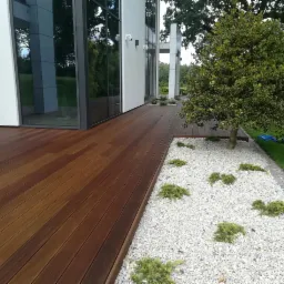 Nowoczesny taras drewniany z desek w kolorze ciemnego brązu przylegający do białego budynku z dużymi oknami, obok rabata z białych kamieni i niskich krzewów.