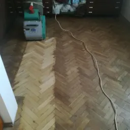 Fragment pokoju z parkietem w jodełkę, częściowo odnowionym szlifierką podłogową Deva, widoczny kabel zasilający, w tle komoda i szafa.