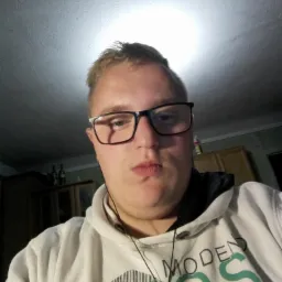 Autoportret mężczyzny w okularach, w białej bluzie z napisem 'Modern Chaos', z perspektywy żabiej, oświetlonego od góry.