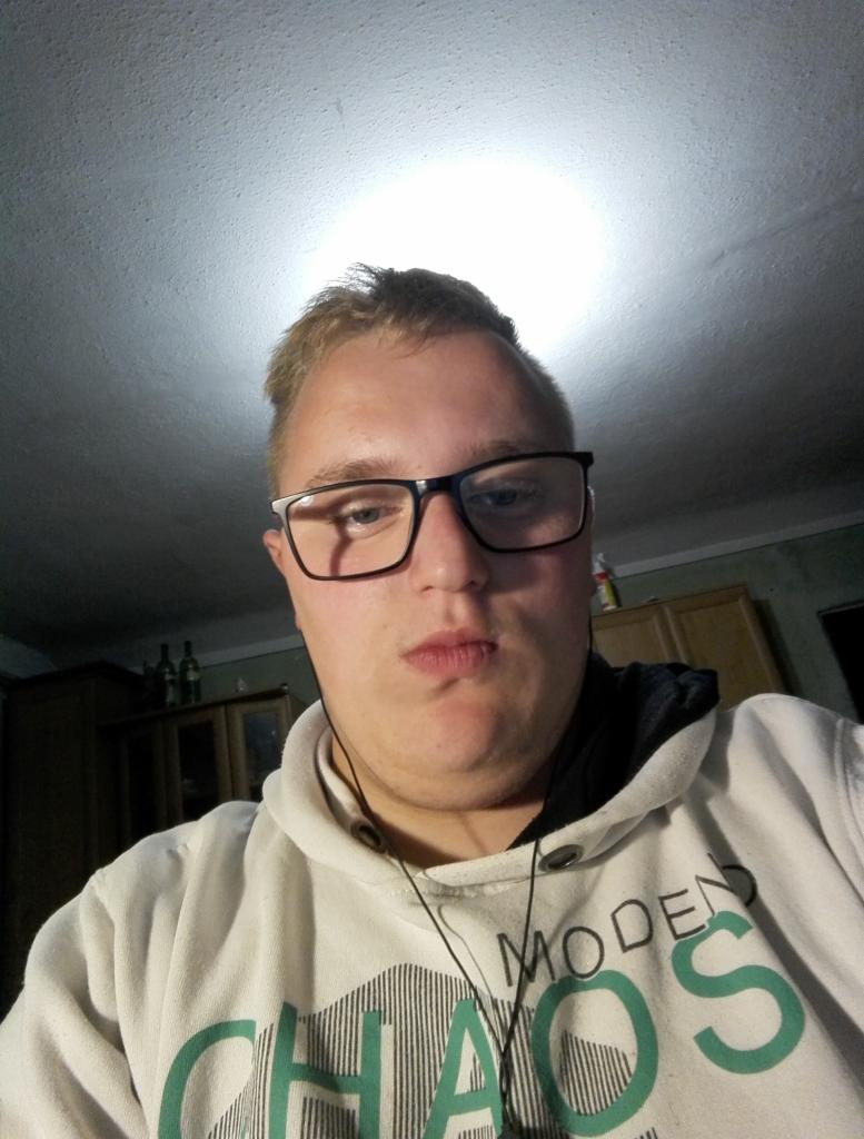 Autoportret mężczyzny w okularach, w białej bluzie z napisem 'Modern Chaos', z perspektywy żabiej, oświetlonego od góry.