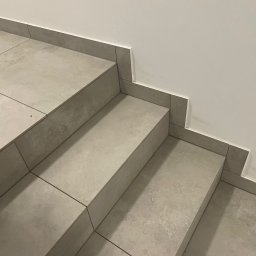 Supra Mateusz Kuśnierz - Wykończone schody z szarych, matowych płytek ceramicznych z fugą w dopasowanym kolorze, z widocznym fragmentem buta w lewym dolnym rogu i śladami po kleju na ścianie.