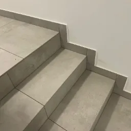 Wykończone schody z szarych, matowych płytek ceramicznych z fugą w dopasowanym kolorze, z widocznym fragmentem buta w lewym dolnym rogu i śladami po kleju na ścianie.