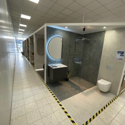 Supra Mateusz Kuśnierz - Nowoczesna ekspozycja łazienki z kabiną prysznicową, umywalką z podświetlanym lustrem i toaletą. Aranżacja w szarościach, z akcentem roślinnym i oświetleniem punktowym.
