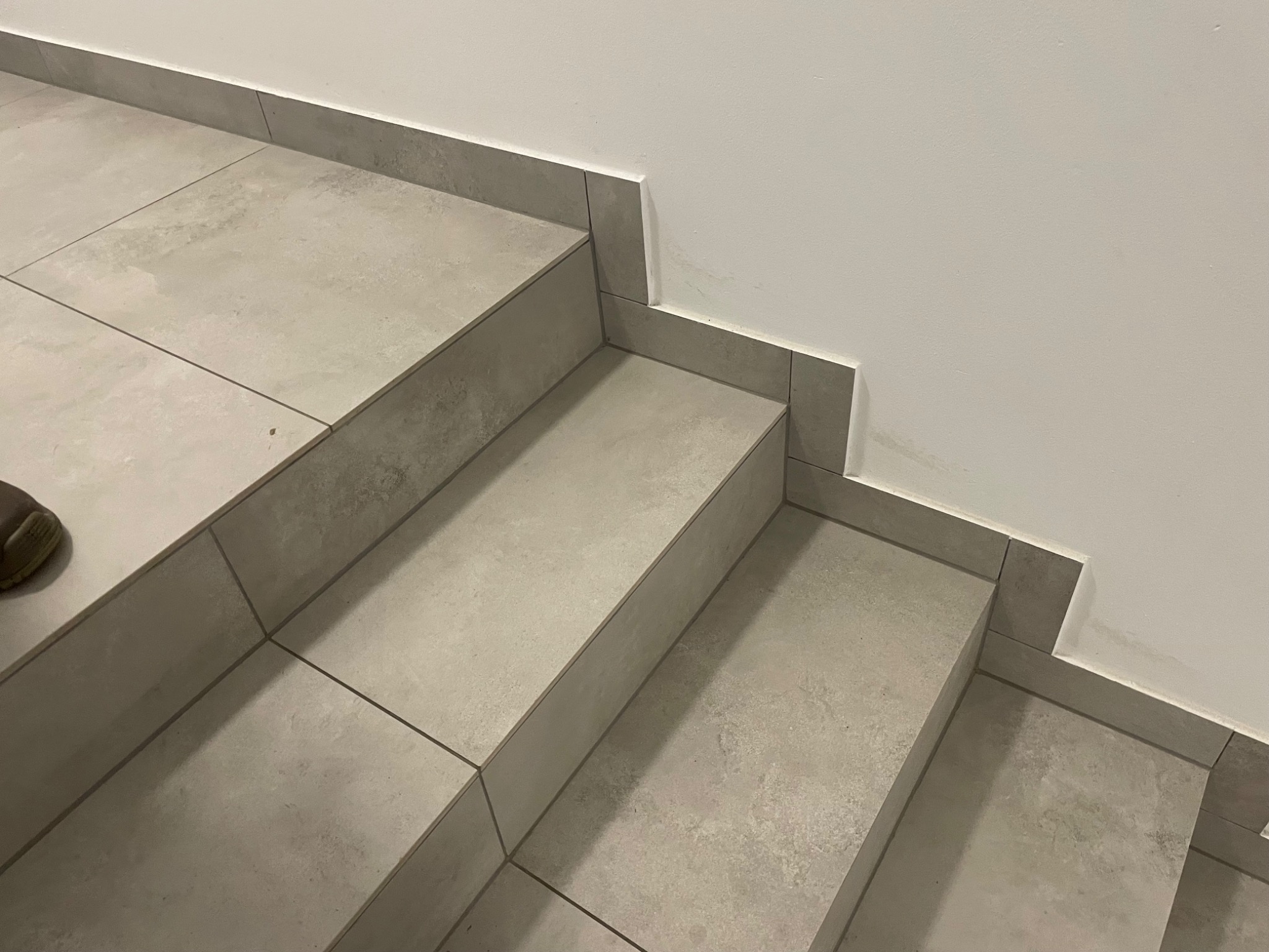 Wykończone schody z szarych, matowych płytek ceramicznych z fugą w dopasowanym kolorze, z widocznym fragmentem buta w lewym dolnym rogu i śladami po kleju na ścianie.