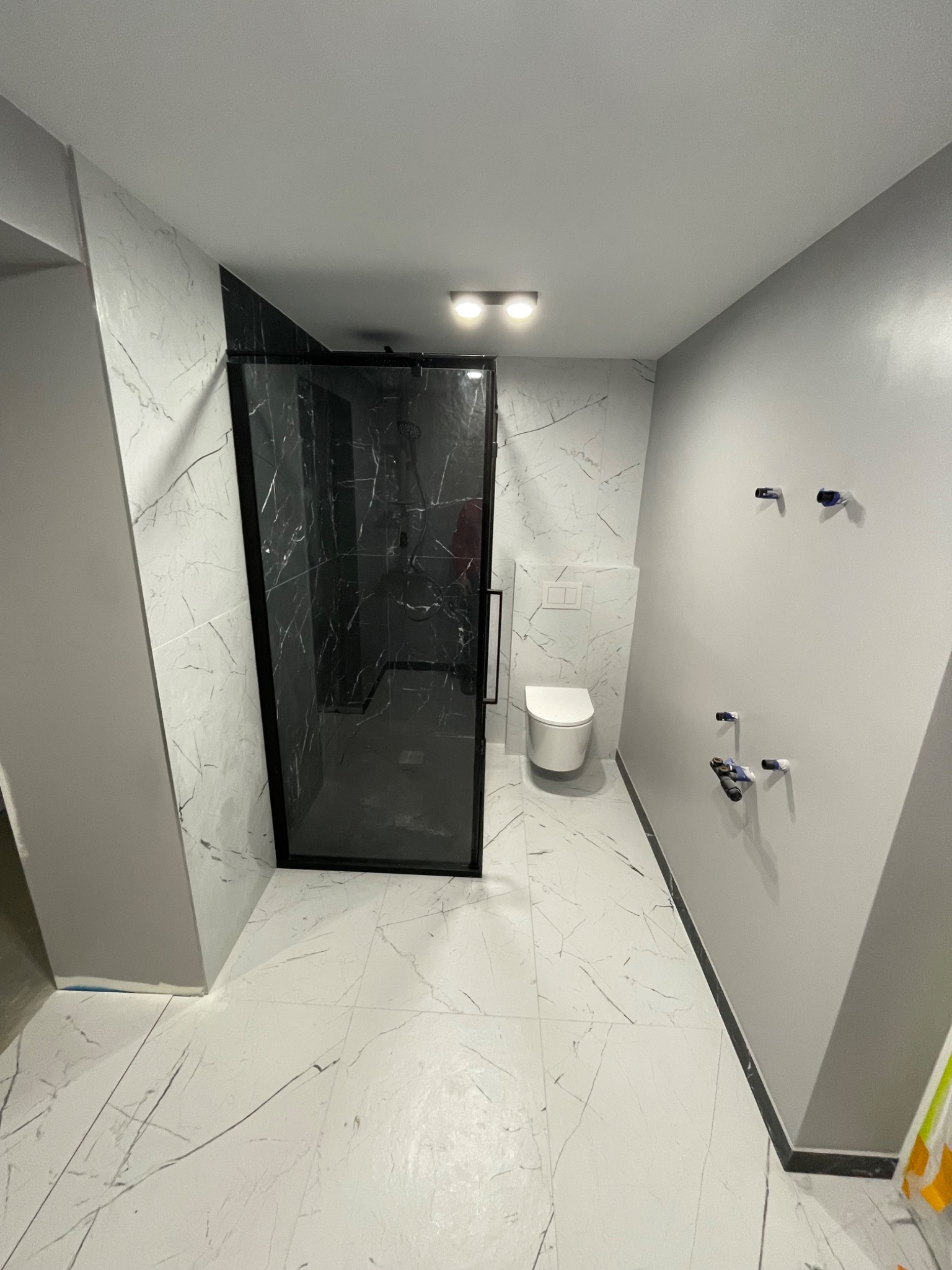 Nowoczesna łazienka z kabiną prysznicową typu walk-in z czarną ramą, białe płytki imitujące marmur na podłodze i ścianach, szara ściana z widocznymi przyłączami wodno-kanalizacyjnymi,...