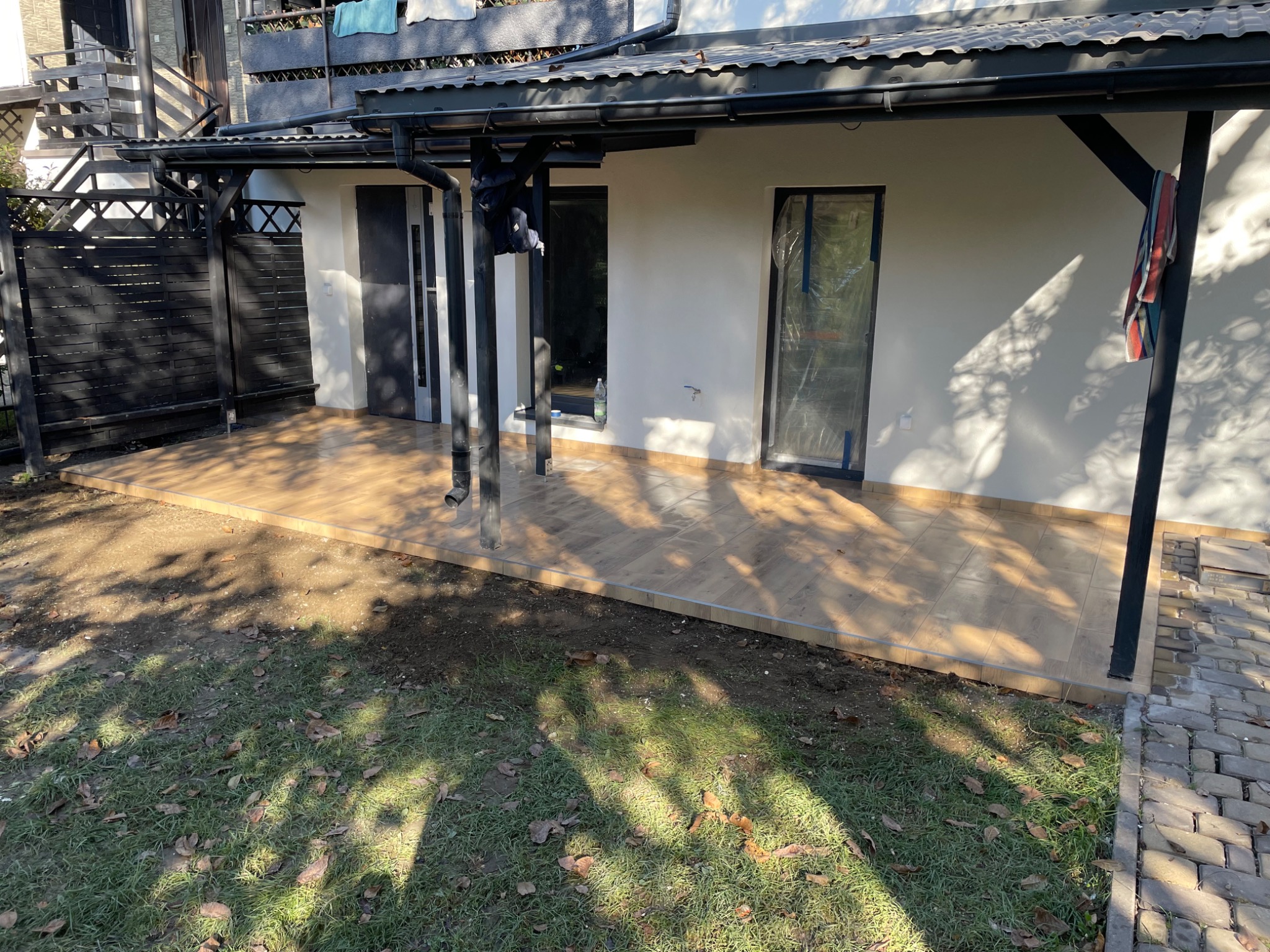 Wyłożony płytkami taras przy budynku z oknami zabezpieczonymi folią malarską, widoczny fragment trawnika z liśćmi.