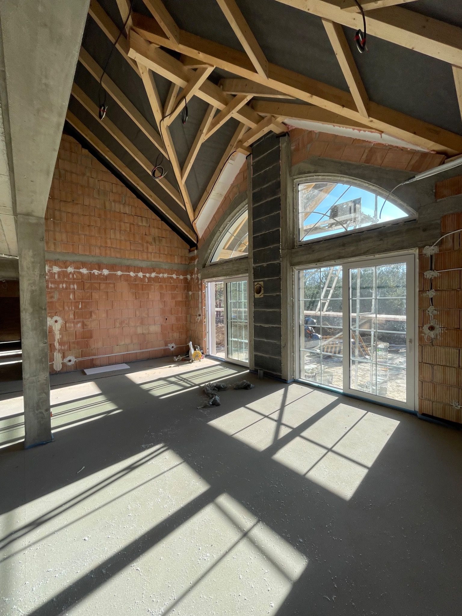 Surowy stan wnętrza domu w budowie: widoczne ceglane ściany z instalacją elektryczną, drewniana konstrukcja dachu i wylewka podłogowa z promieniami słońca.