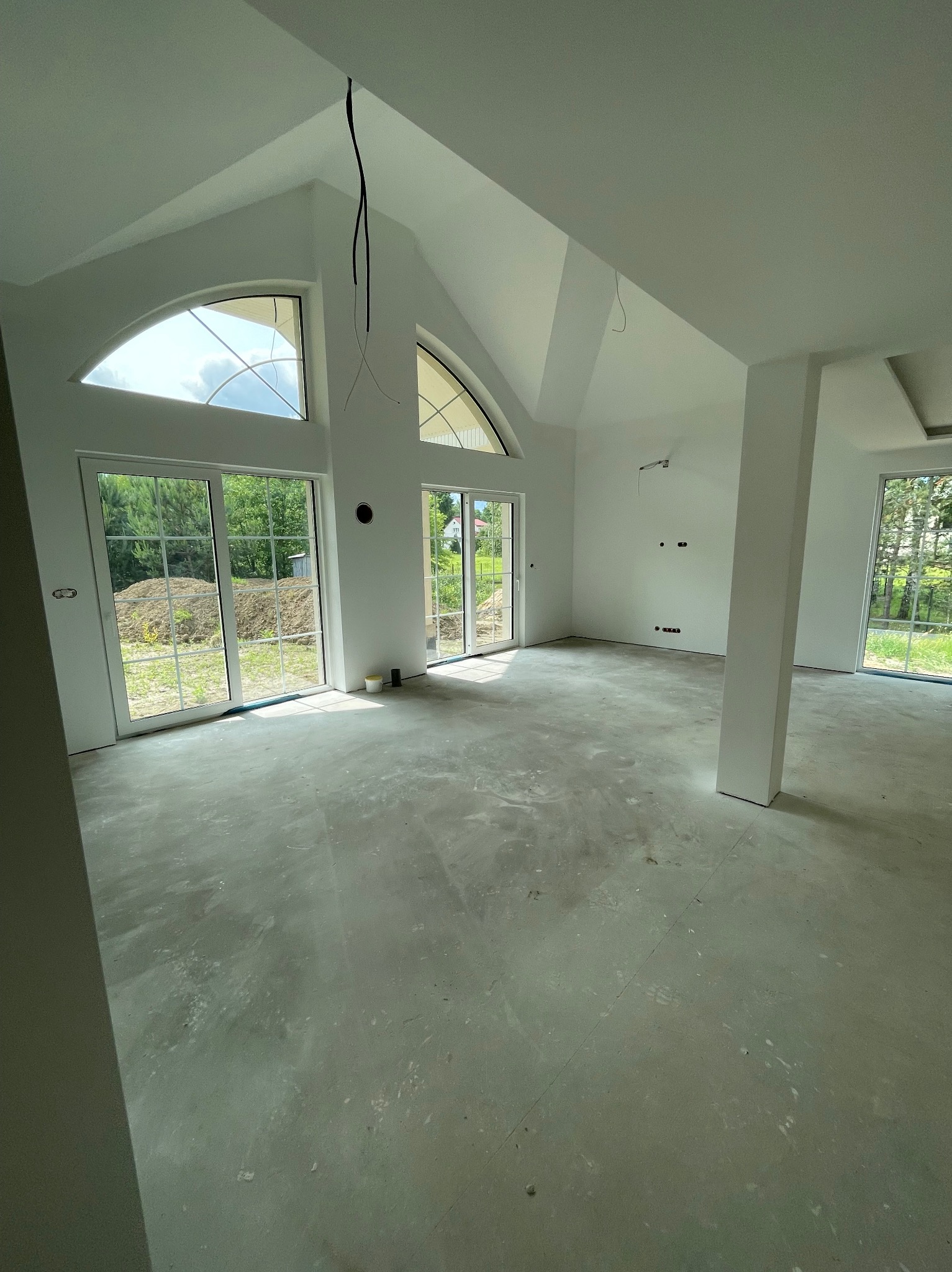 Surowe wnętrze domu z dużymi oknami i wysokim sufitem. Widoczne przewody elektryczne, ściany pomalowane na biało, surowa podłoga. Dom w stanie deweloperskim.