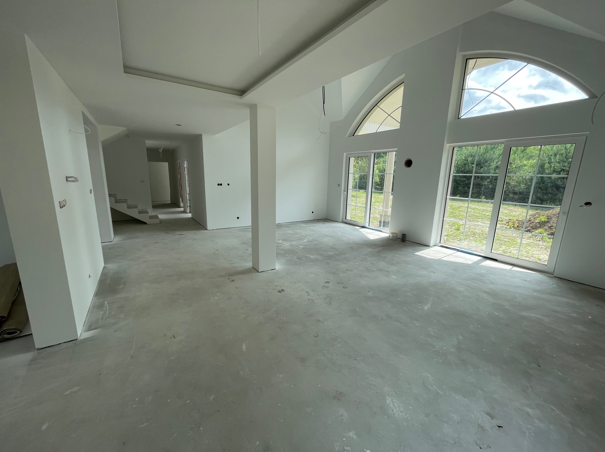 Białe, przestronne wnętrze domu w stanie deweloperskim z dużymi oknami i widokiem na zieleń. Ściany pomalowane na biało, surowa podłoga, widoczne instalacje elektryczne.