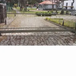 Otwarta, przesuwna brama automatyczna z szarego metalu na posesji z kostki brukowej, widok z zewnątrz.