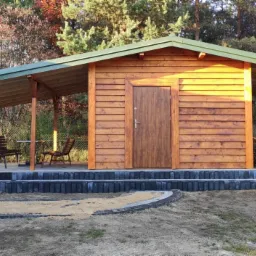 Drewniana altana z zadaszeniem i schodami z kostki brukowej, obok ułożone drewno na opał, w tle drzewa.