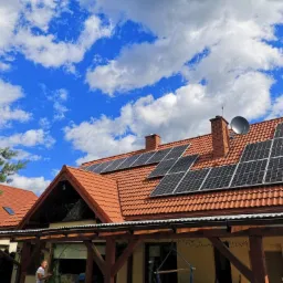 Czerwony dach z dachówki ceramicznej, panele słoneczne na połaci, antena satelitarna na kominie, błękitne niebo z obłokami w tle.