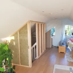 Poddasze w trakcie adaptacji: drewniana konstrukcja ścianki działowej wypełniona wełną mineralną, widoczne schody, podłoga z paneli, w tle okno i fragment umeblowania.