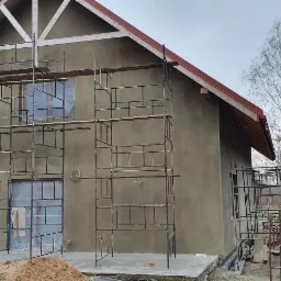 Dom jednorodzinny w trakcie prac elewacyjnych, widoczne rusztowania i zabezpieczone okna folią, świeżo położony tynk w kolorze szaro-beżowym, przygotowany grunt pod elewację.