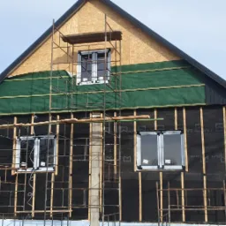 Dom w trakcie ocieplania: widoczna czarna membrana izolacyjna, zielona folia w górnej części ściany, drewniane listwy, nowe okna PCV i rusztowanie.
