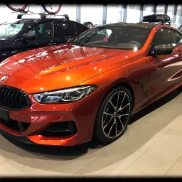 Pomarańczowe BMW serii 8 coupe z czarnymi felgami i dachem, stojące w salonie samochodowym, z innymi autami w tle.