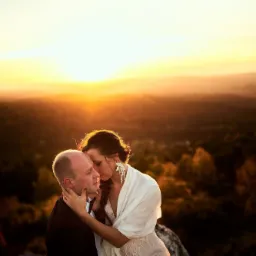 Para młoda w objęciach na tle zachodzącego słońca, ona w białej sukni i szalu, on w ciemnym garniturze, plenerowa sesja z delikatnym oświetleniem.