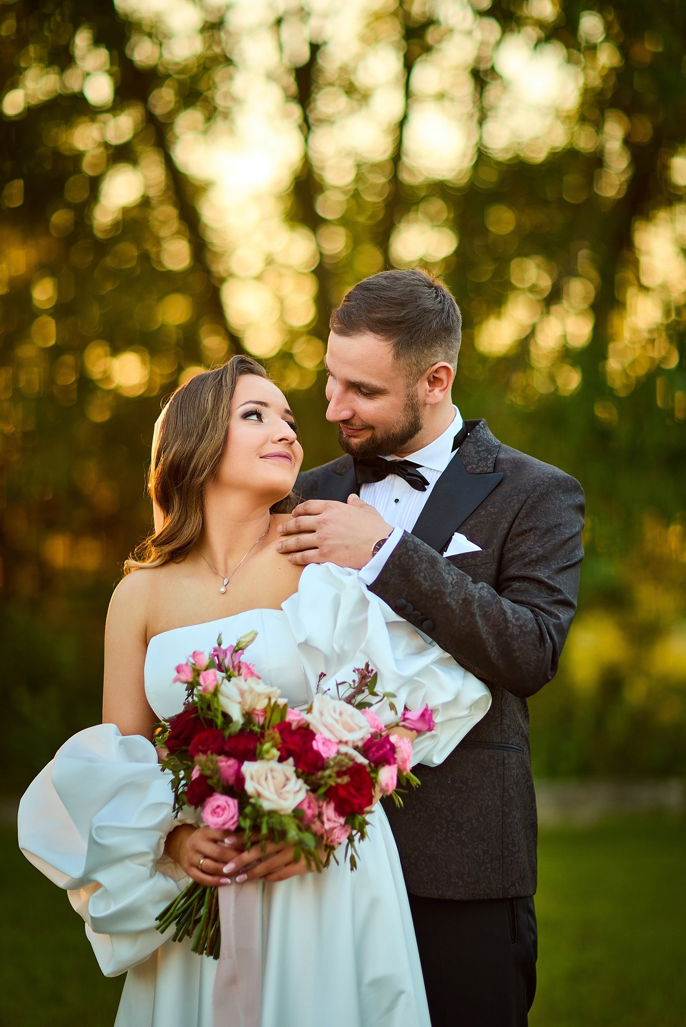 Para młoda w plenerze: panna młoda w sukni z bufiastymi rękawami trzyma bukiet, pan młody w eleganckim smokingu obejmuje ją czule. Romantyczna sceneria, złote światło.