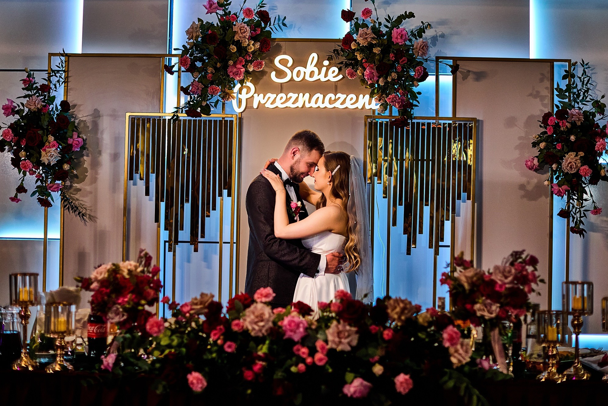 Para młoda w objęciach na tle dekoracji weselnej ze świetlnym napisem 'Sobie Przeznaczeni' i kompozycjami kwiatowymi w odcieniach różu i czerwieni.