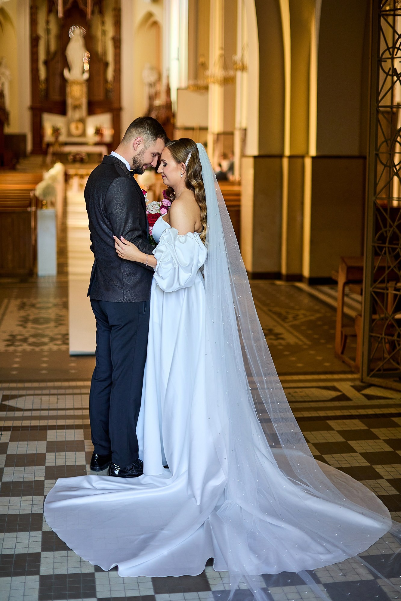 Para młoda w kościele: Panna młoda w białej sukni z długim welonem, Pan młody w czarnym smokingu, delikatny uścisk, intymna atmosfera, dekoracje kościelne w tle.