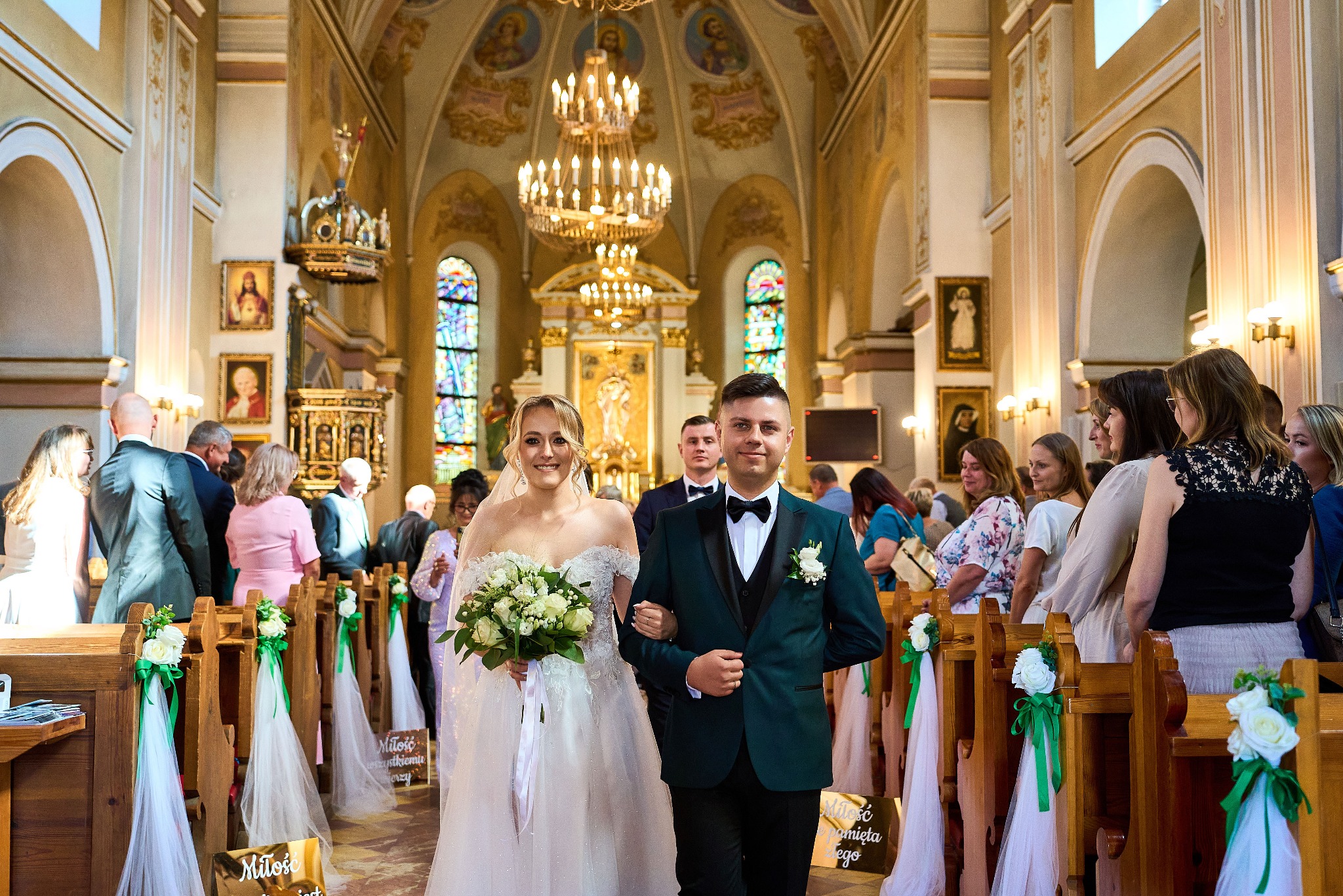 Para młoda w kościele, on w butelkowej zieleni smokingu, ona w białej sukni z bukietem. Dekoracje kwiatowe na ławkach, goście po bokach. W tle ołtarz i witraże.