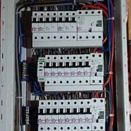 Rozdzielnica elektryczna z zabezpieczeniami różnicowoprądowymi i nadprądowymi, widoczne kable w peszlach wchodzące od góry, zamontowana na ścianie z widoczną izolacją.