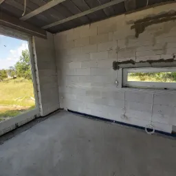 Surowy stan wnętrza budynku w trakcie prac instalacyjnych: widoczne okno z widokiem na zewnątrz, ściany z bloczków, instalacje elektryczne prowadzone po ścianach i pod sufitem, w tle widoczne...