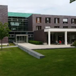 Nowoczesny budynek kliniki rehabilitacyjnej z zielonym dachem, widoczny pacjent na wózku inwalidzkim w towarzystwie opiekuna na ścieżce prowadzącej do wejścia.