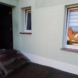 Świeżo pomalowana elewacja budynku z dwoma oknami, z których jedno jest uchylone, widoczne schody wejściowe i fragment chodnika z kostki brukowej.