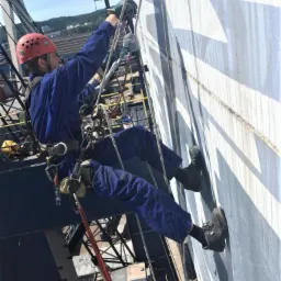 Pracownik w kombinezonie i kasku, zabezpieczony linami, maluje zewnętrzną ścianę zbiornika przemysłowego na wysokości, z widocznymi zaciekami świeżej farby.