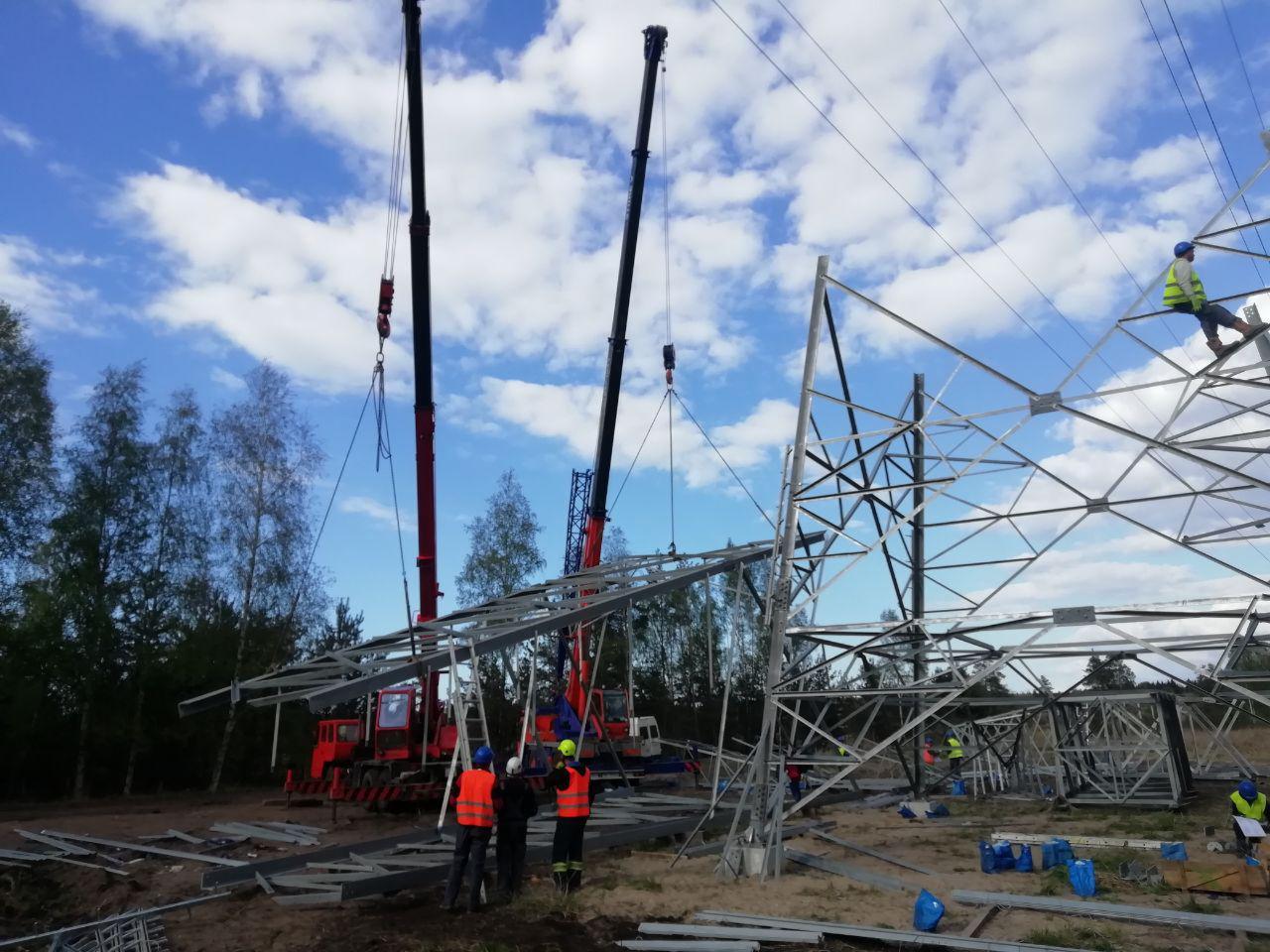 Montaż stalowej konstrukcji słupa wysokiego napięcia przy użyciu dwóch dźwigów, pracownicy w odblaskowych kamizelkach i kaskach na placu budowy, błękitne niebo z chmurami w tle.