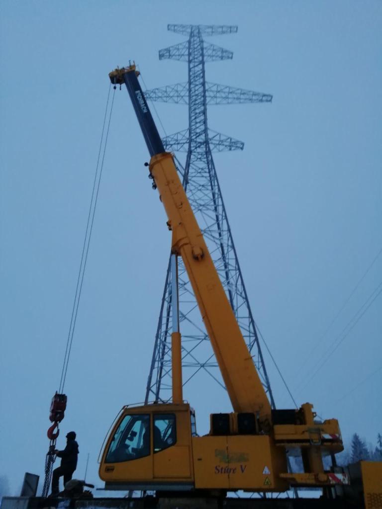 Żółty dźwig teleskopowy z operatorem na tle stalowej konstrukcji słupa wysokiego napięcia w pochmurny dzień.