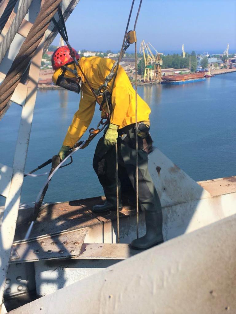 Pracownik w żółtym kombinezonie i czerwonym kasku, zabezpieczony linami, czyści metalową konstrukcję mostu za pomocą myjki ciśnieniowej; w tle widok na port i morze.