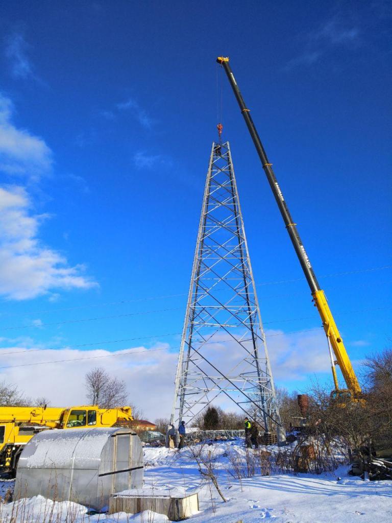 Montaż stalowej wieży energetycznej z użyciem dwóch dźwigów teleskopowych na tle zimowego krajobrazu i błękitnego nieba.