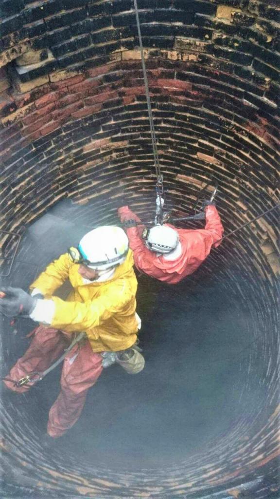 Dwóch pracowników w kaskach i kombinezonach, na linach, wewnątrz ceglanego komina podczas czyszczenia lub inspekcji.