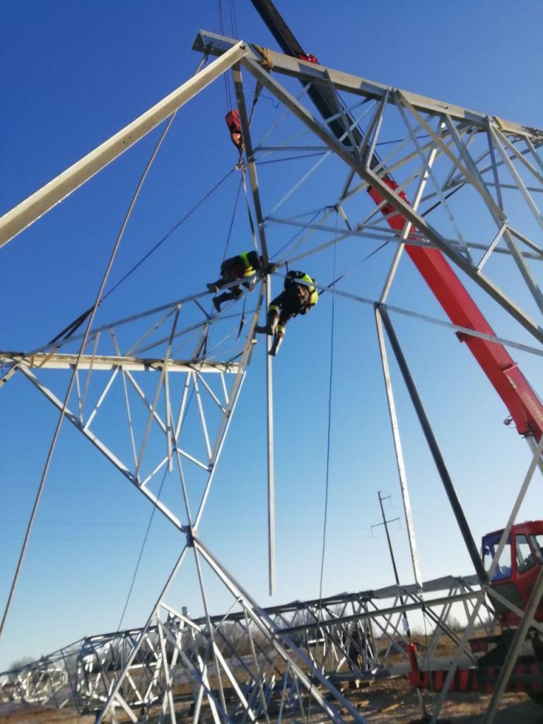 Montaż stalowej konstrukcji słupa wysokiego napięcia przez dwóch pracowników w uprzężach, z użyciem dźwigu