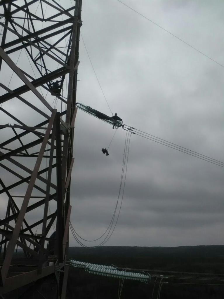 Elektryk pracujący na słupie wysokiego napięcia na tle pochmurnego nieba, widoczne elementy konstrukcji słupa i izolatory.