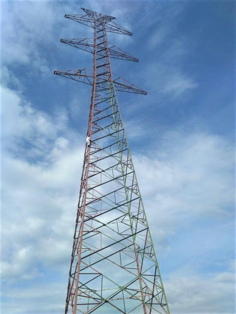 Wysoka wieża energetyczna pomalowana częściowo na czerwono i zielono, z pracownikiem w białym kombinezonie widocznym na jednej z poprzeczek, na tle błękitnego nieba z chmurami.